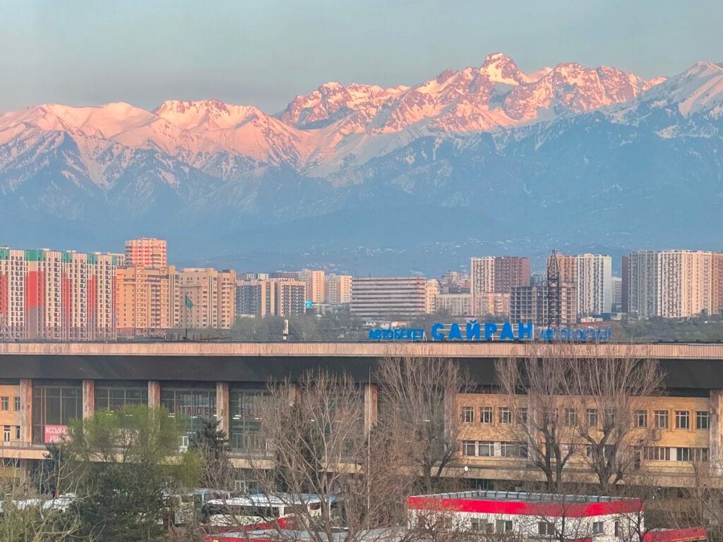 Almaty cityscape with the snow-capped Zailiyskiy Alatau mountains glowing pink at sunset, viewed from a nearby apartment
