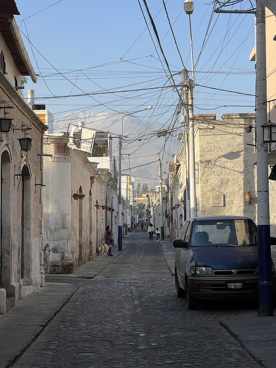 Quiet cobblestone street in Arequipa, Peru, lined with white volcanic stone buildings and crisscrossed power lines, with hazy mountains in the distance.