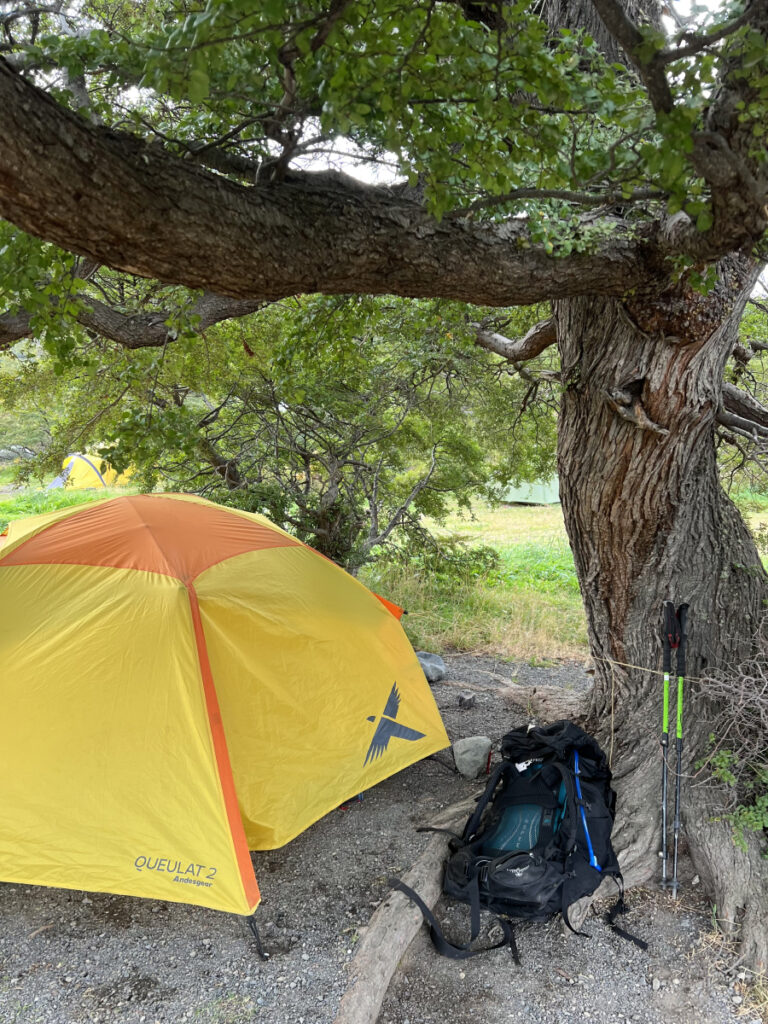 Yellow and orange Queulat tent pitched under a tree with a backpack and trekking poles on the ground — a typical campsite on the O Circuit in Torres del Paine National Park, Patagonia