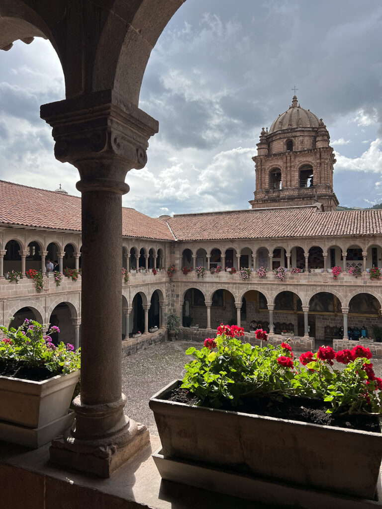 Colonial courtyard at the Convent of Santo Domingo in Cusco, Peru, with stone arches, flower boxes, and a domed bell tower under a cloudy sky.