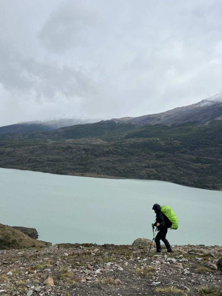 Hiker with a neon green rain cover ascending a rocky trail above a turquoise lake on the O Circuit in Torres del Paine, Patagonia, under cloudy skies
