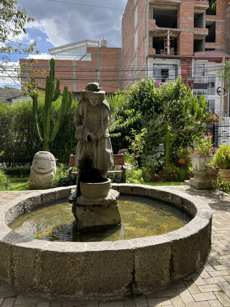 Stone fountain with traditional statue in a museum courtyard garden in Huaraz, Peru, framed by cactus plants and urban buildings in the background.