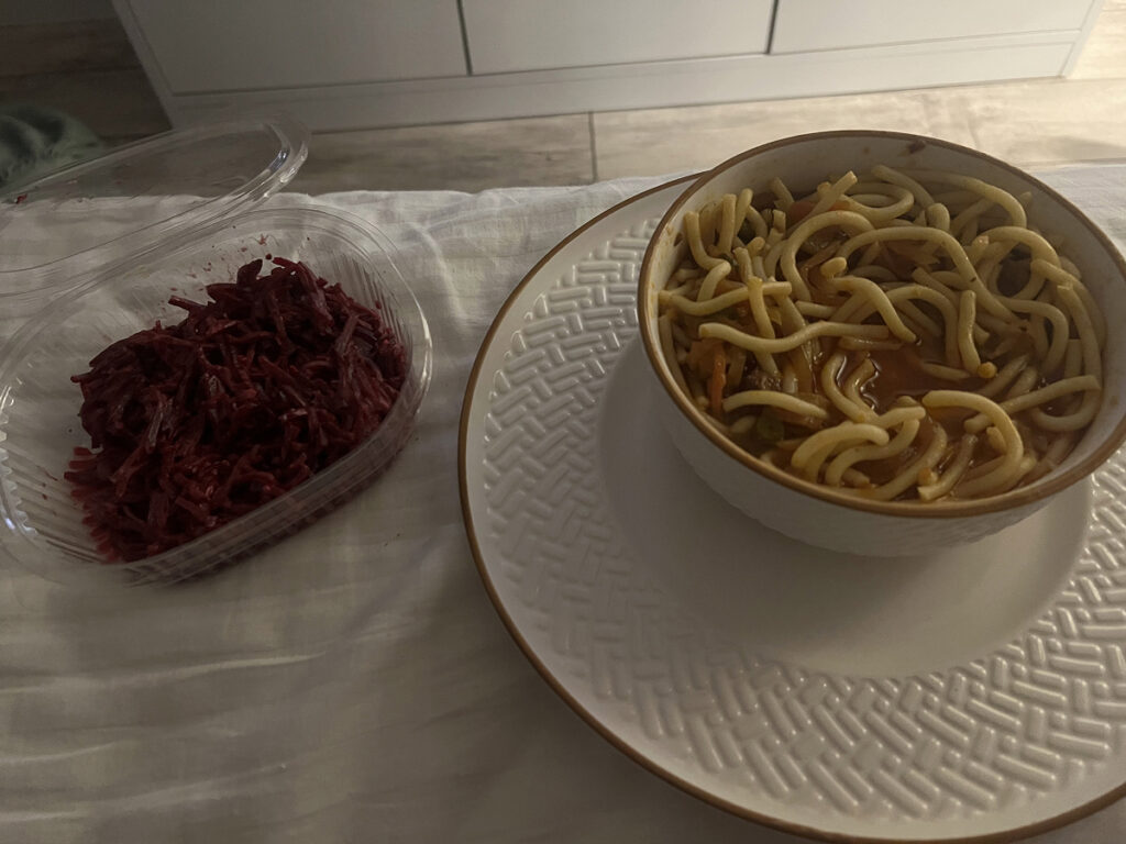 Bowl of laghman noodles in broth with a side of beet salad, served on a bed during a rainy night in Almaty