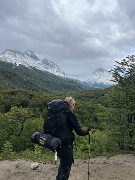 Backpacker standing with trekking poles on the O Circuit in Torres del Paine, overlooking a lush green valley and dramatic snow-capped mountains under cloudy skies in Patagonia