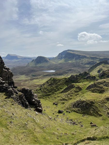 Isle of Skye hiking scenery from the Quiraing trail, showing dramatic green hills, layered rock formations, and a distant loch