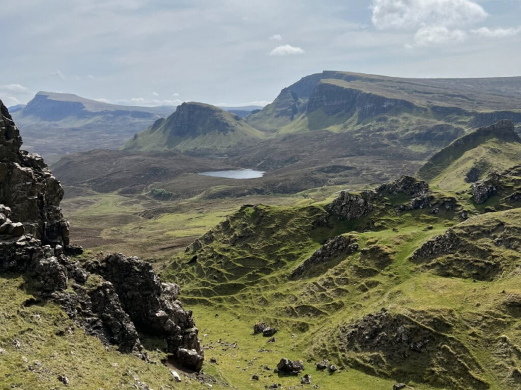 View from the Quiraing hike on the Isle of Skye, showing dramatic green hills, layered rock formations, and a small lake in the distance under a partly cloudy sky