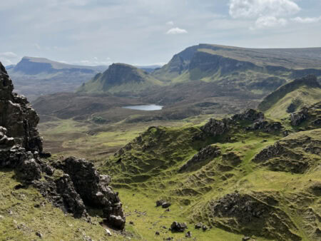 View from the Quiraing hike on the Isle of Skye, showing dramatic green hills, layered rock formations, and a small lake in the distance under a partly cloudy sky