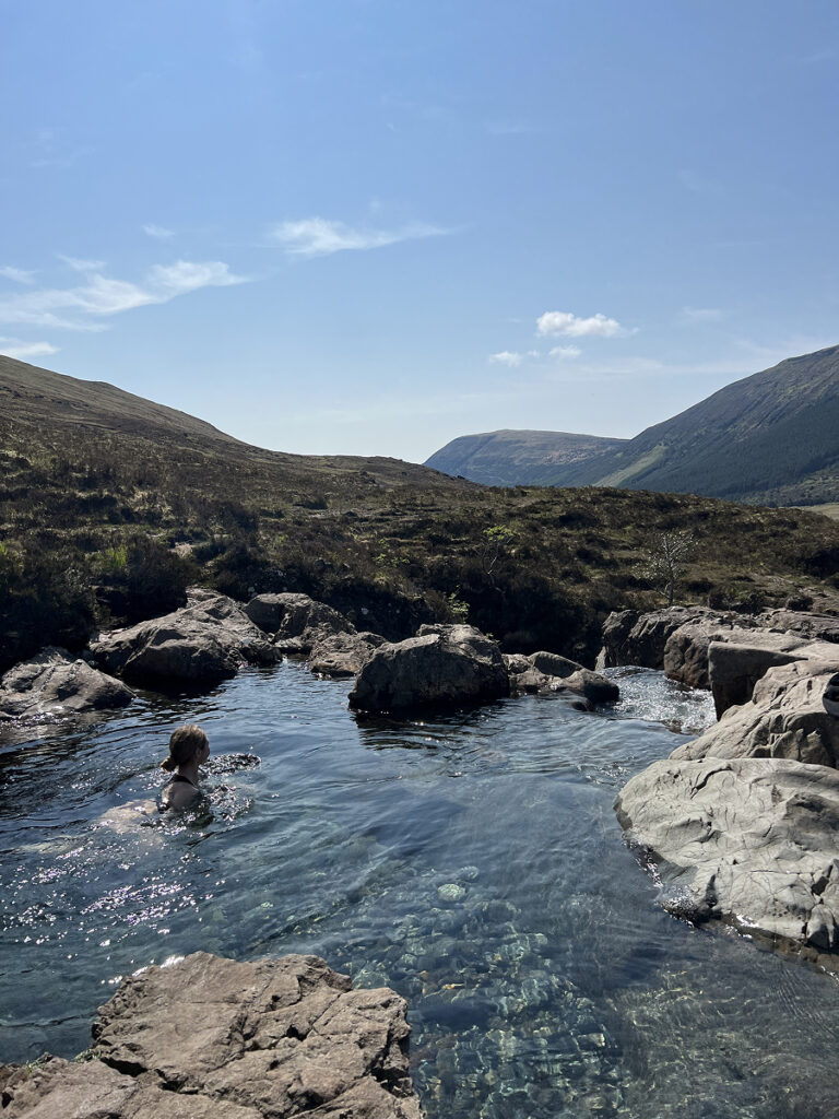 Woman swimming in a crystal-clear pool surrounded by rocks on the Fairy Pools trail, under a blue sky in the Scottish Highlands