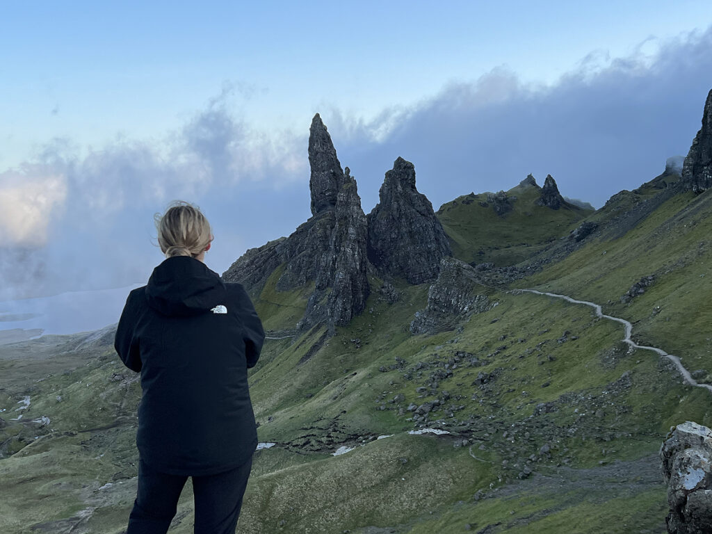 Hiker in black jacket standing at a viewpoint facing the Old Man of Storr rock pinnacles on the Isle of Skye during the evening, with mist rolling in