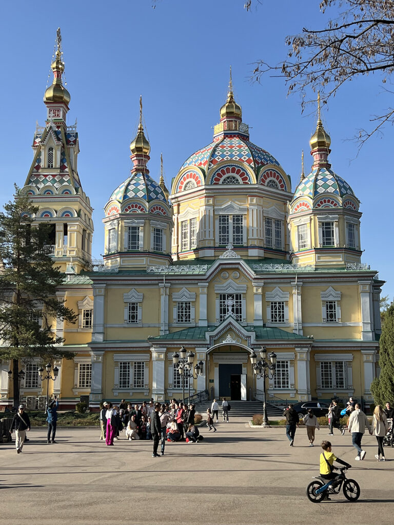 Front view of Zenkov Cathedral in Almaty, Kazakhstan, with its colorful domes, ornate wooden facade, and people walking in front on a sunny day