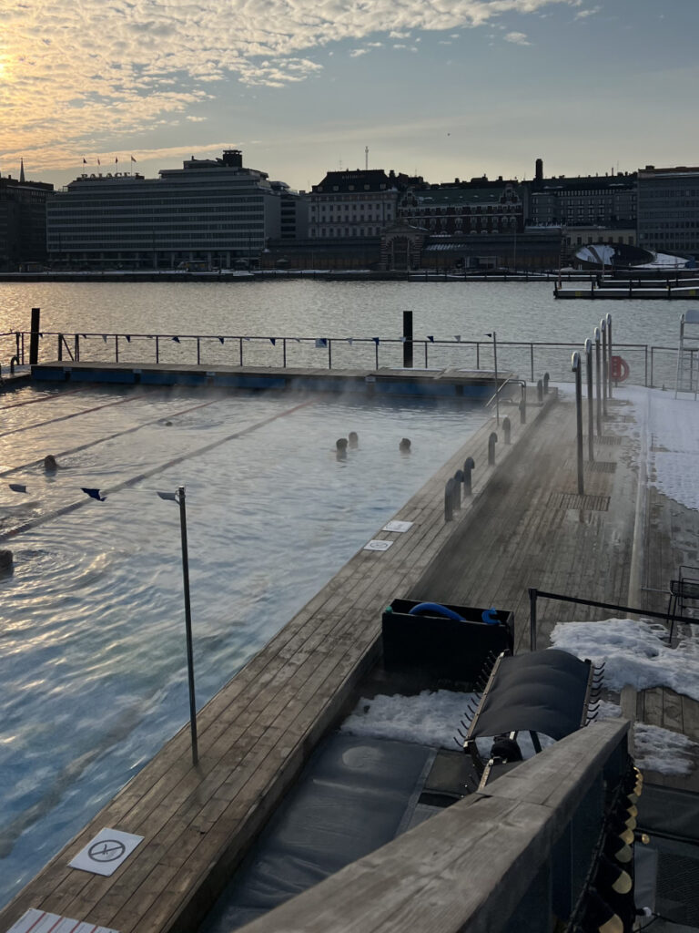 Winter view of Allas Sea Pool in Helsinki, with steam rising from the warm pool and the harbor skyline in the background at sunset.