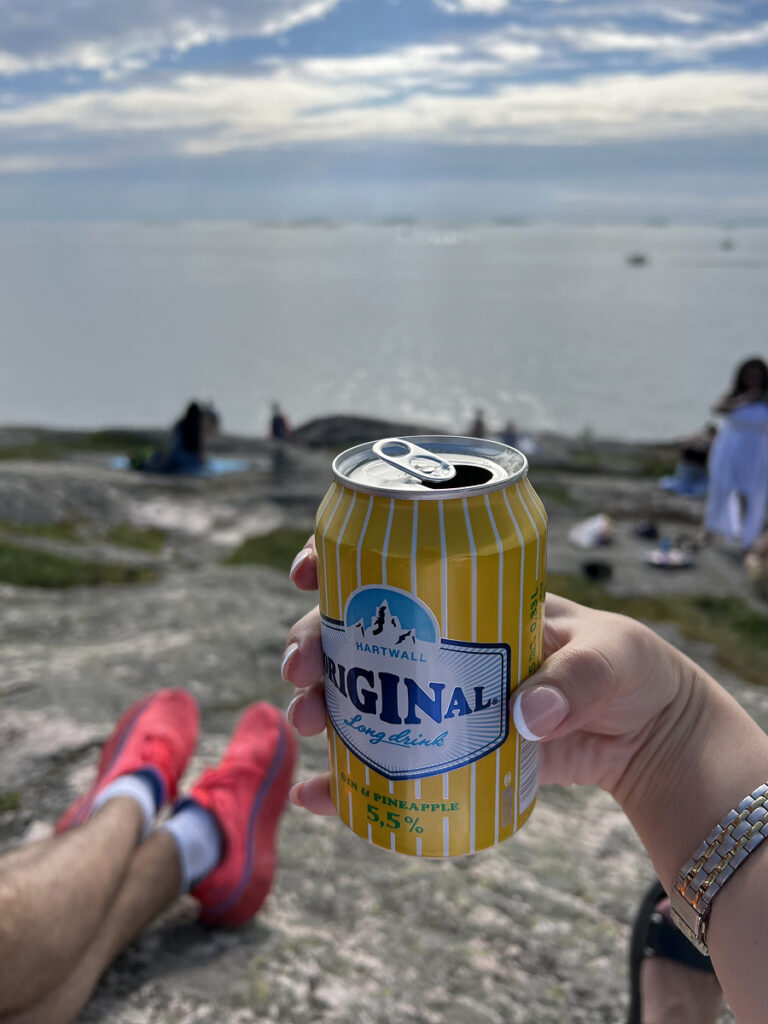 Holding a can of Hartwall Original Long Drink at Suomenlinna Fortress Island in Helsinki, with sea views in the background.