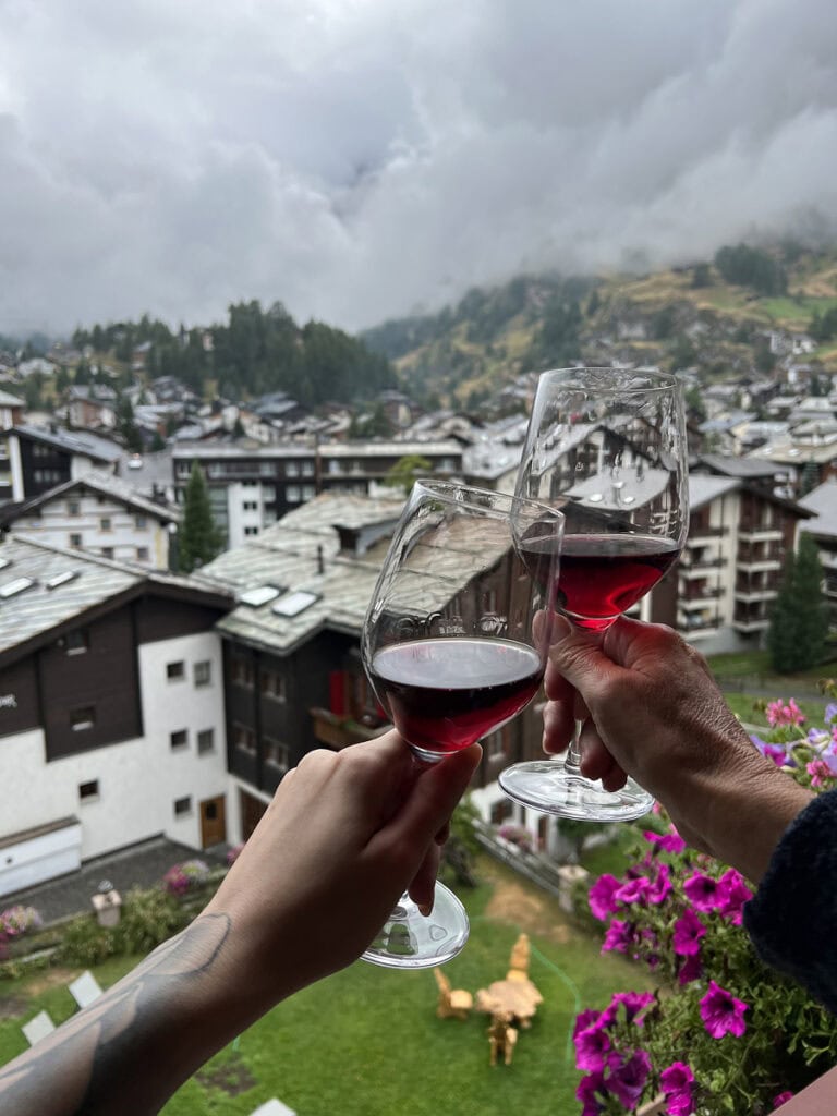 Toasting glasses of red wine on the balcony at Capricorn Hotel Zermatt with views over the village rooftops.