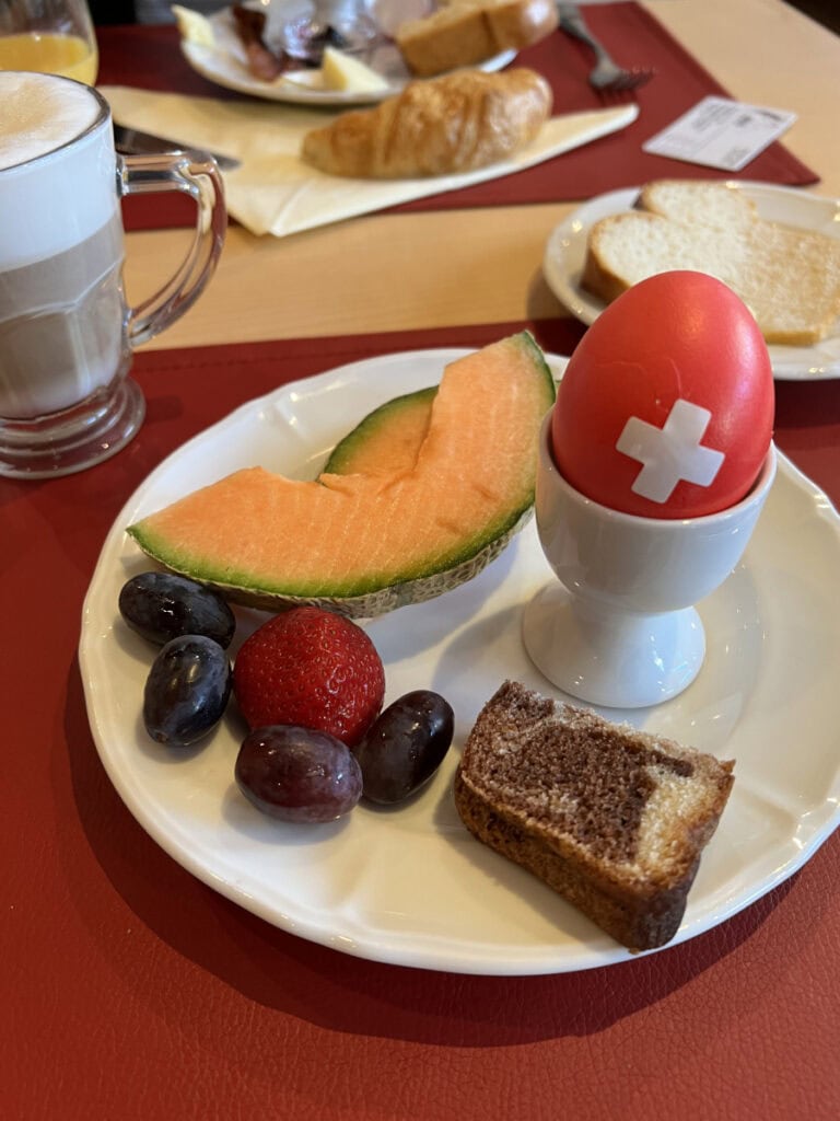 Hotel Capricorn Zermatt breakfast plate with red Swiss cross egg, fruit, pastry, and cappuccino.