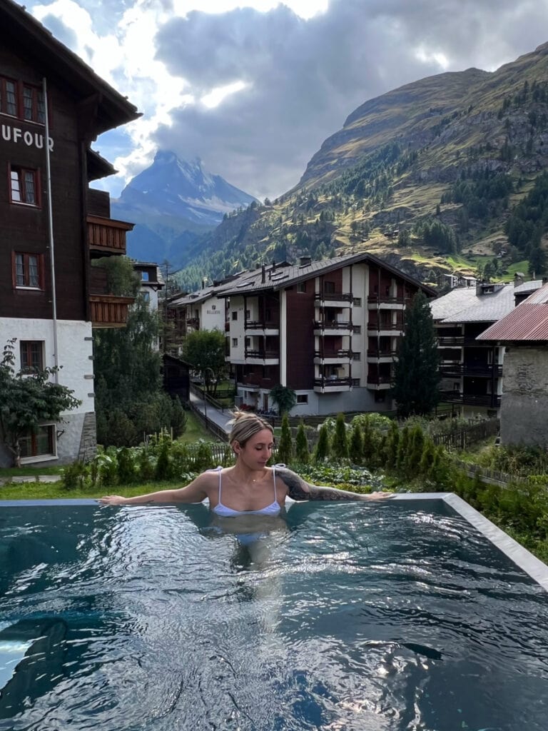 Guest in the outdoor pool at Hotel Capricorn Zermatt with mountain views in the background.