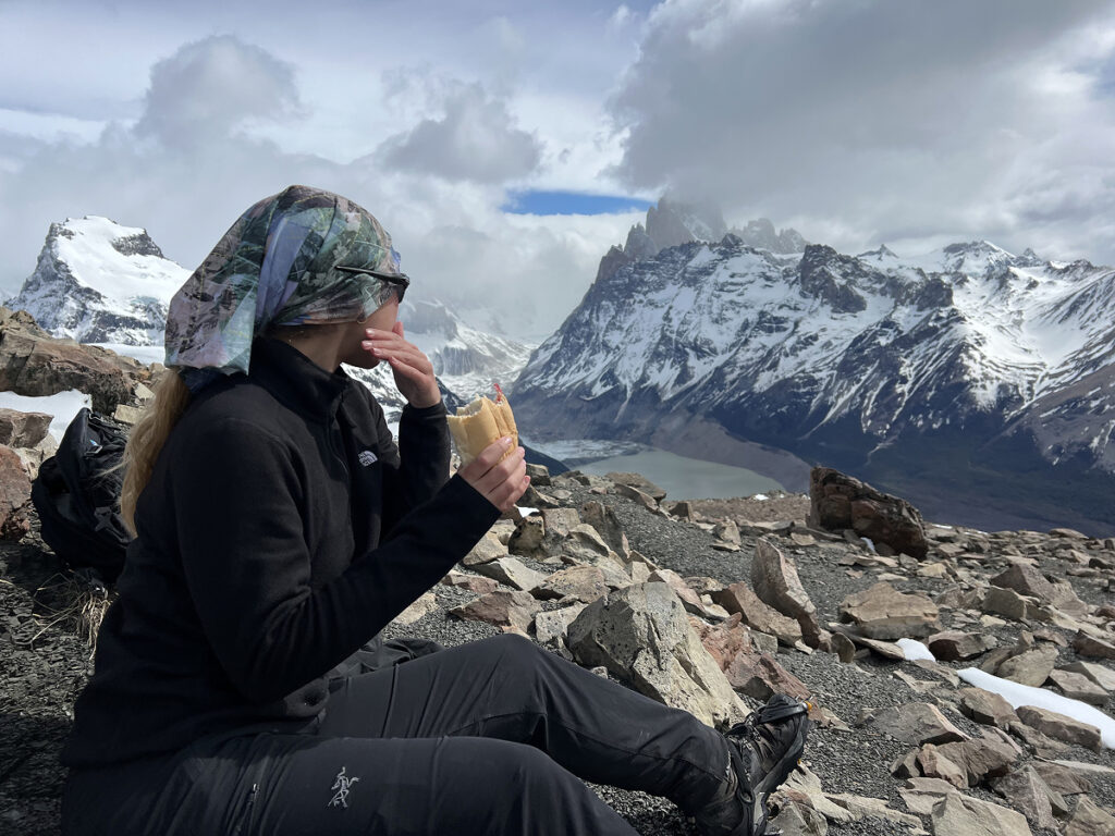 Woman eating a sandwich with panoramic mountain views at the top of Loma del Pliegue Tumbado hike in Patagonia