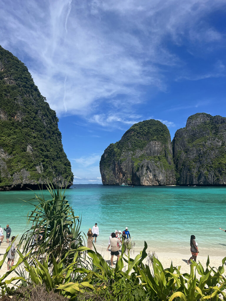 Tourists on the white sand beach of Maya Bay, Phi Phi Islands, Thailand, with dramatic limestone cliffs and turquoise water in the background.