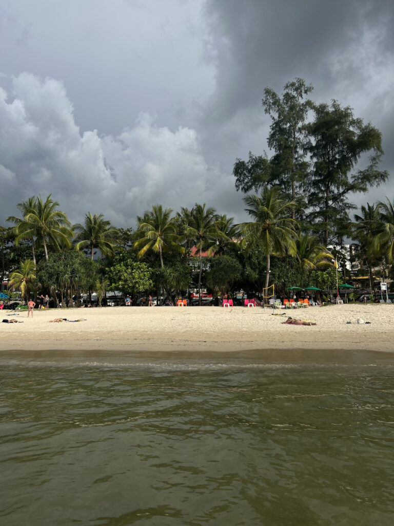 Cloudy sky over the beach in Phuket City, Thailand, with palm trees and sunbathers lining the shore.